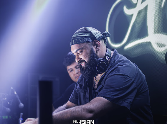DJ with headphones and beard, focused on mixing, in a dimly lit club with a neon sign in the background.