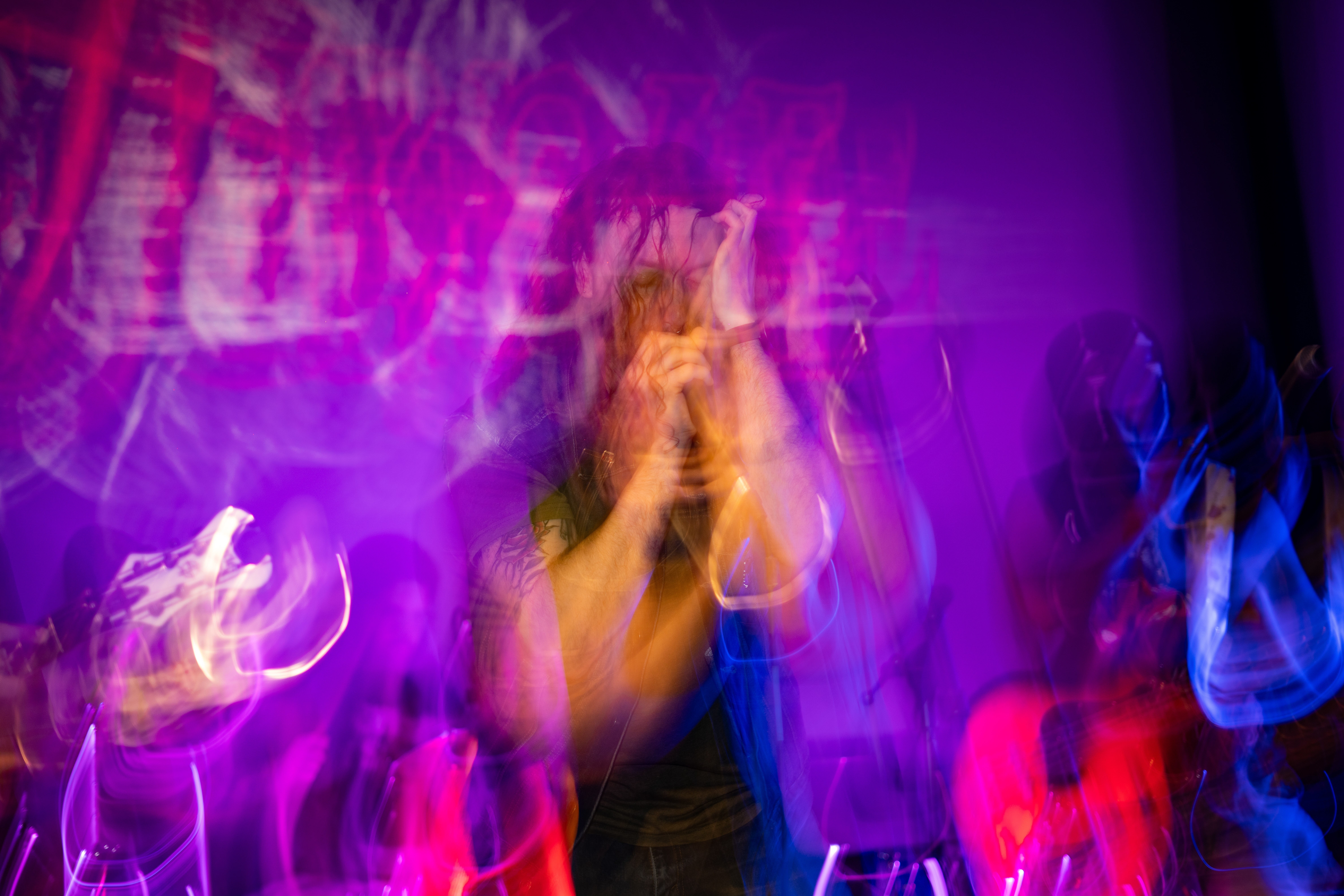 Person with hands near face in a colorful, blurry, vibrant setting with purple and blue lights.