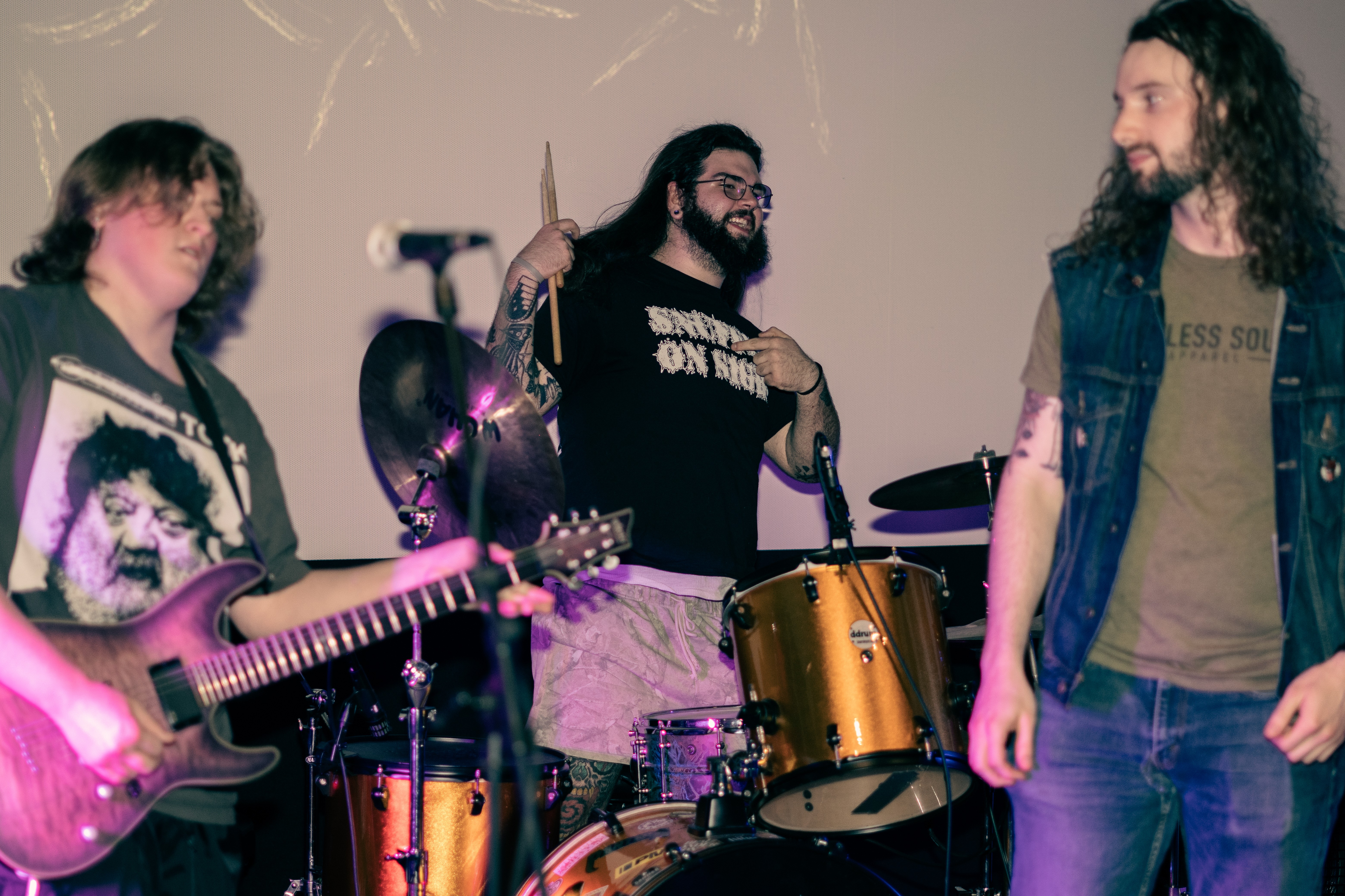 Three musicians perform on stage, one playing guitar, one on drums, and one standing, smiling, with a microphone nearby.