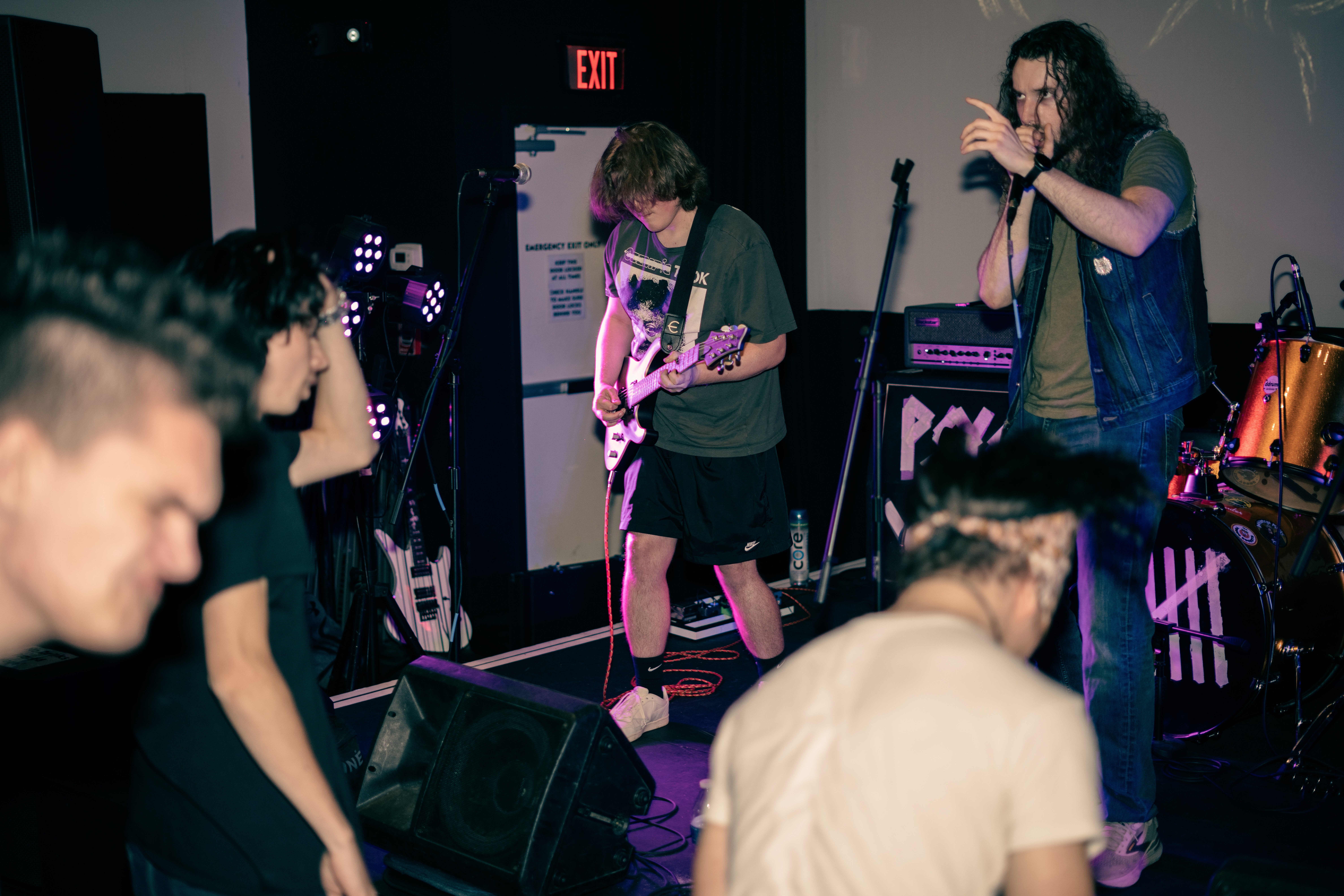 Two musicians perform on stage with audience members watching, one playing guitar and the other singing into a microphone.