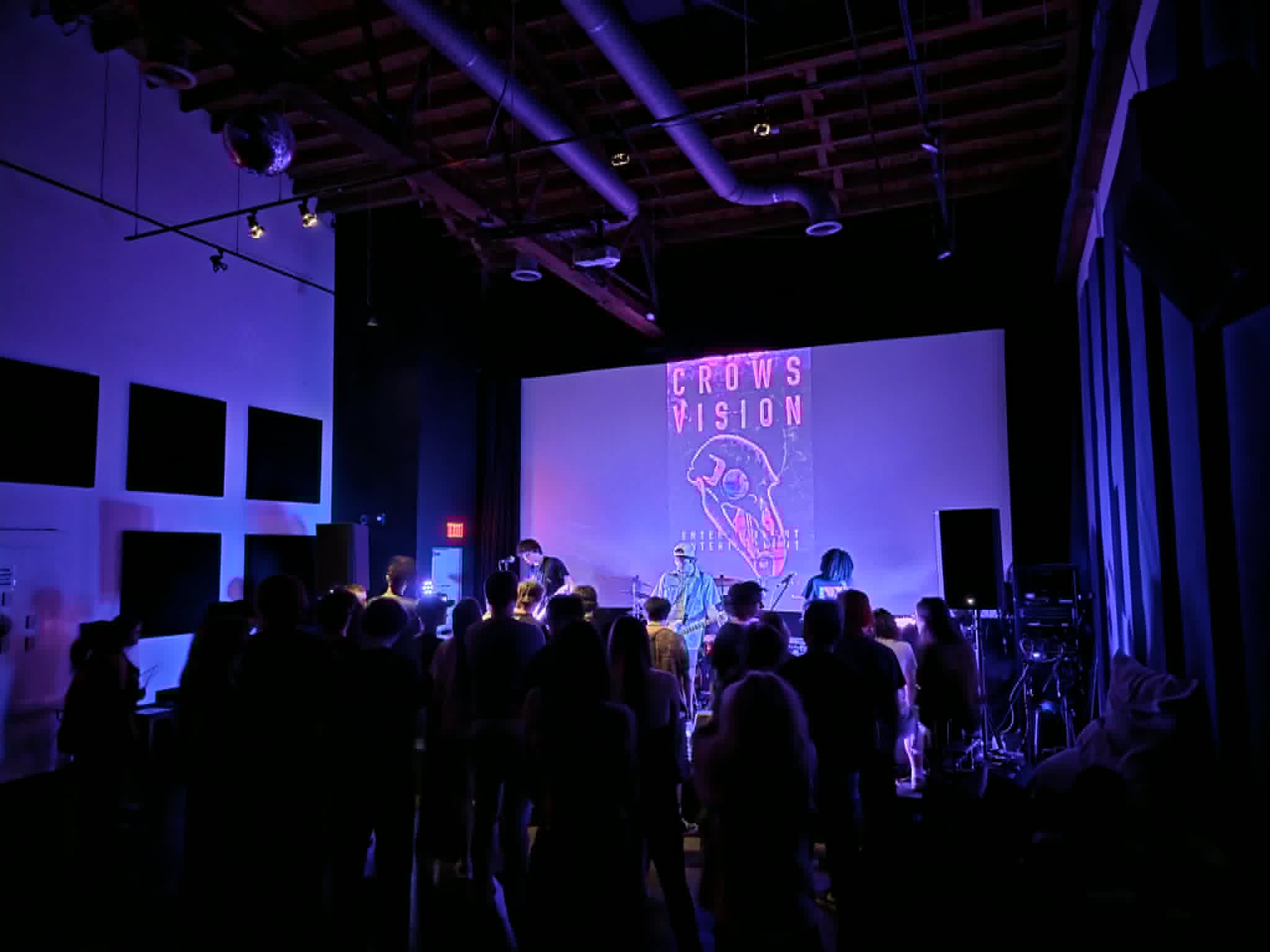 People gather in a dark venue with a stage, purple lighting, and a large screen displaying a logo and text.