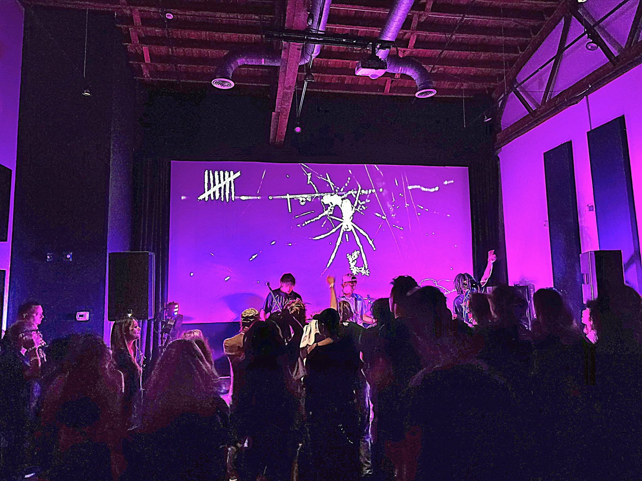 People gathered in a dark room watching a performance on a large purple screen with white lines and splashes.