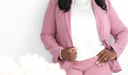 Woman in pink suit smiling, standing with hands on hips, white background with pink text, blurred flowers in foreground.
