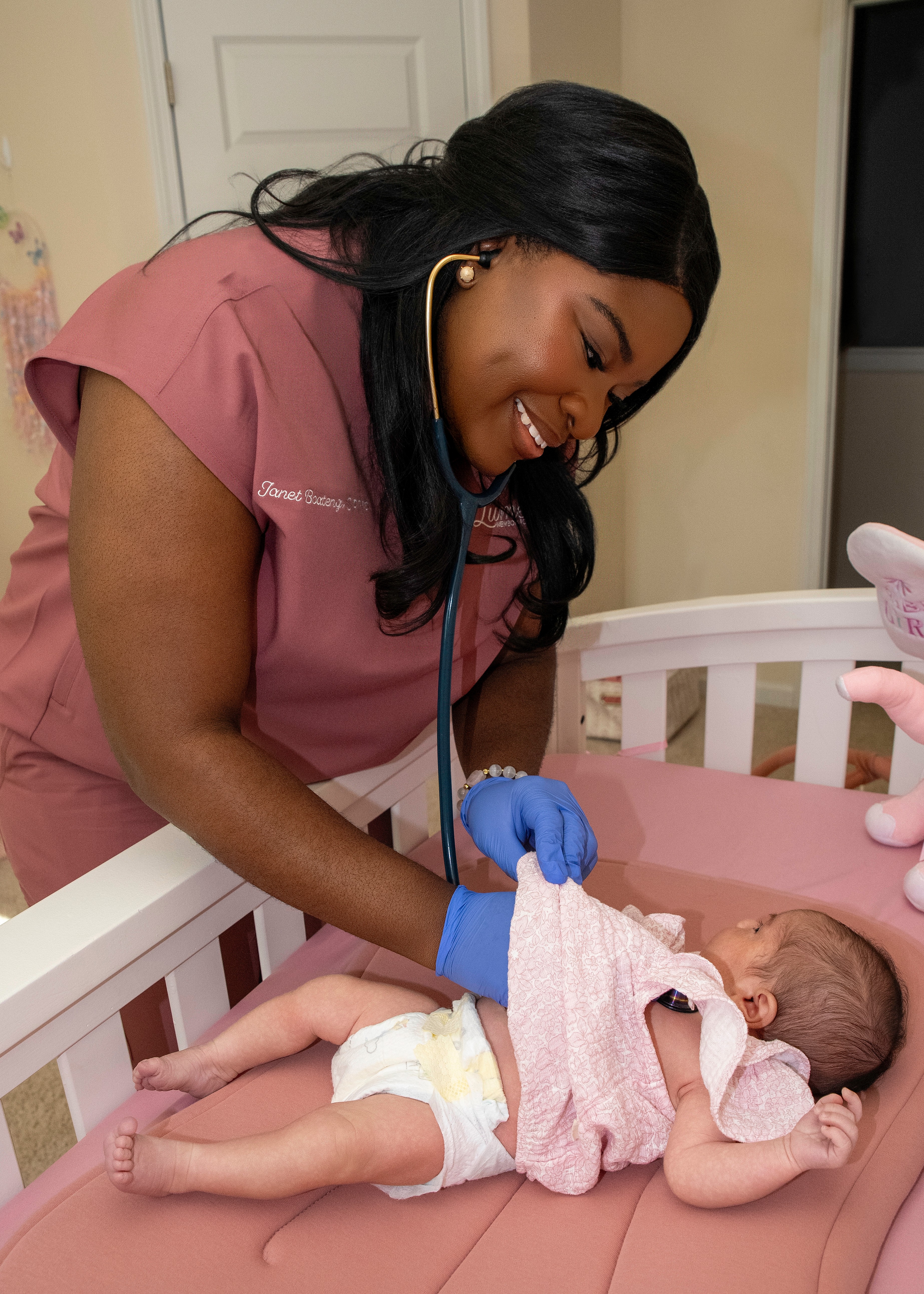 A nurse in pink scrubs and blue gloves gently holds a newborn baby in a crib, smiling warmly.