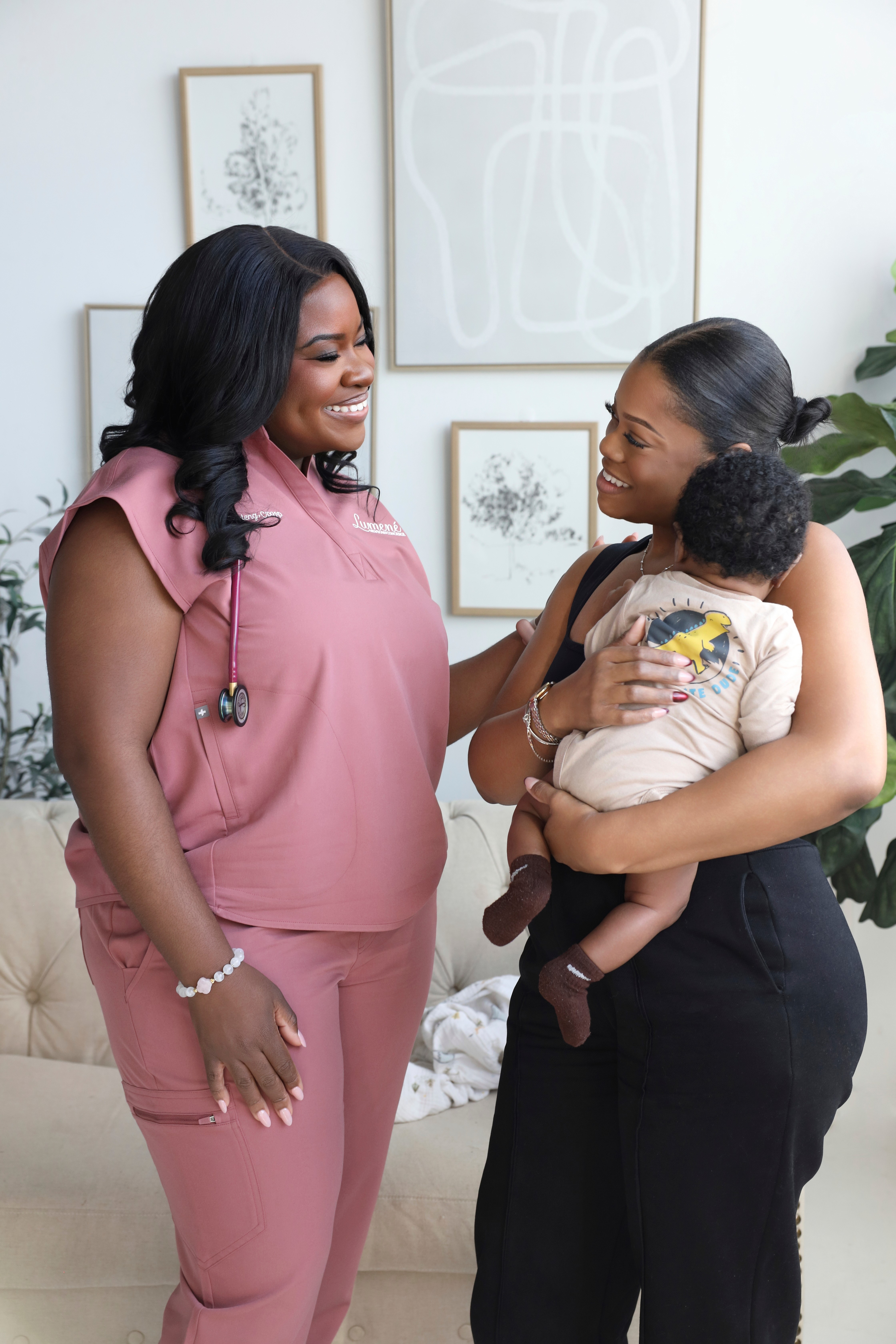 Two women, one holding a child, smiling and talking indoors with framed artwork on the wall behind them.