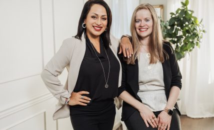 Two women standing together indoors, smiling, with one woman resting her arm on the other's shoulder, in front of a white wall and plant.