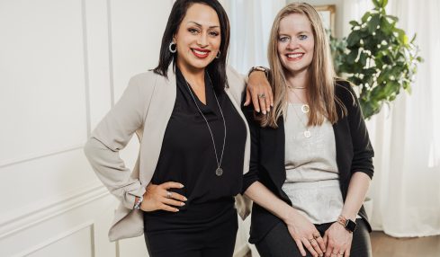 Two women standing together indoors, smiling, with one woman resting her arm on the other's shoulder, in front of a white wall and plant.