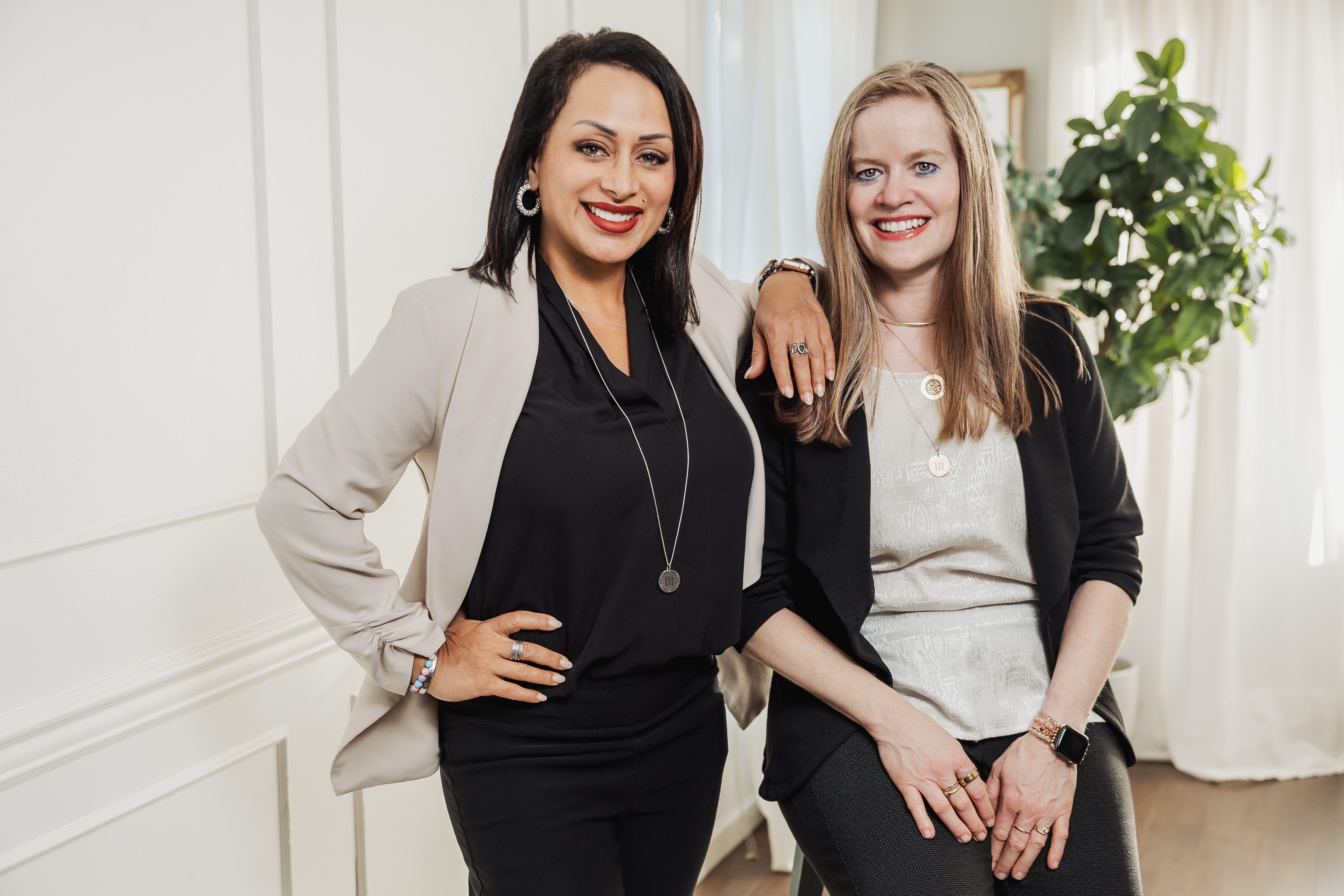 Two women standing together indoors, smiling, with one woman resting her arm on the other's shoulder, in front of a white wall and plant.