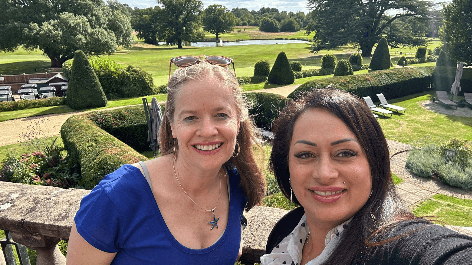 Two women smiling outdoors in a garden with trimmed bushes and trees, sunny weather, and a clear sky.
