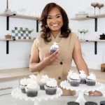 Smiling woman holding cupcake in a bakery with cupcakes on display, shelves with baked goods in background.