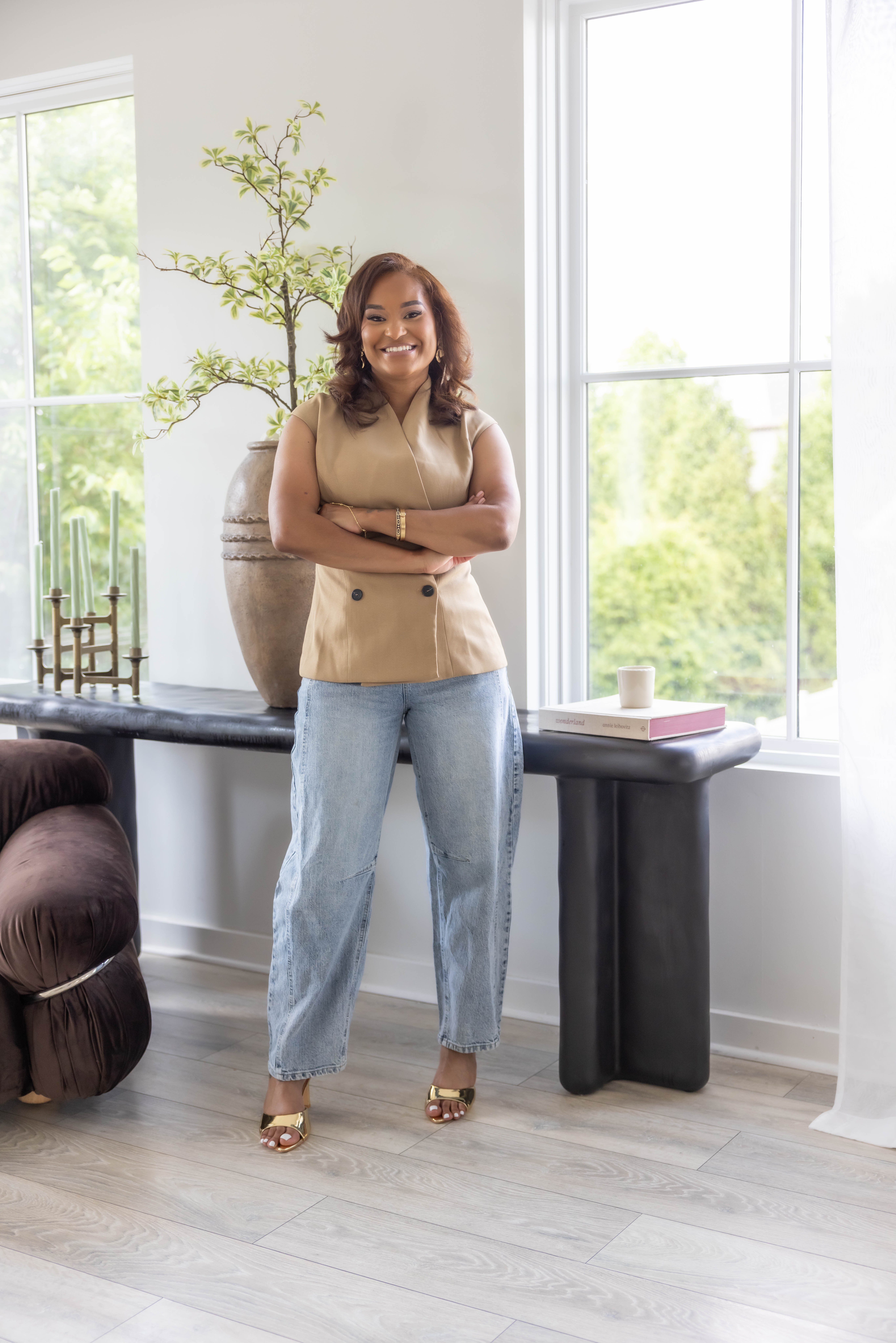 Woman standing indoors near large windows with a potted plant, smiling, wearing a beige top and jeans.