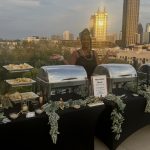 Person standing behind a buffet table with food, city skyline in the background, evening sky, table decorated with greenery.
