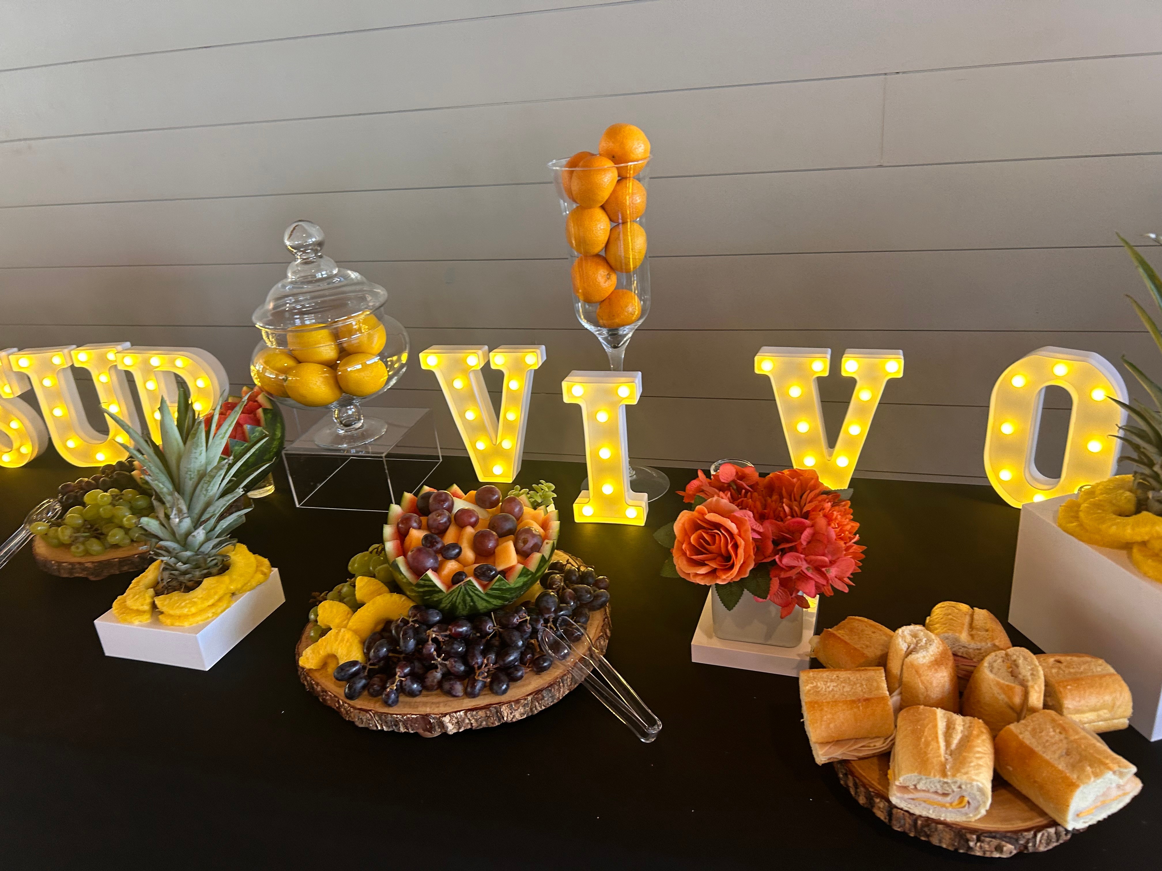 Decorative table with illuminated letters spelling 'VIVO', fruit arrangements, flowers, and desserts, against a white wall background.