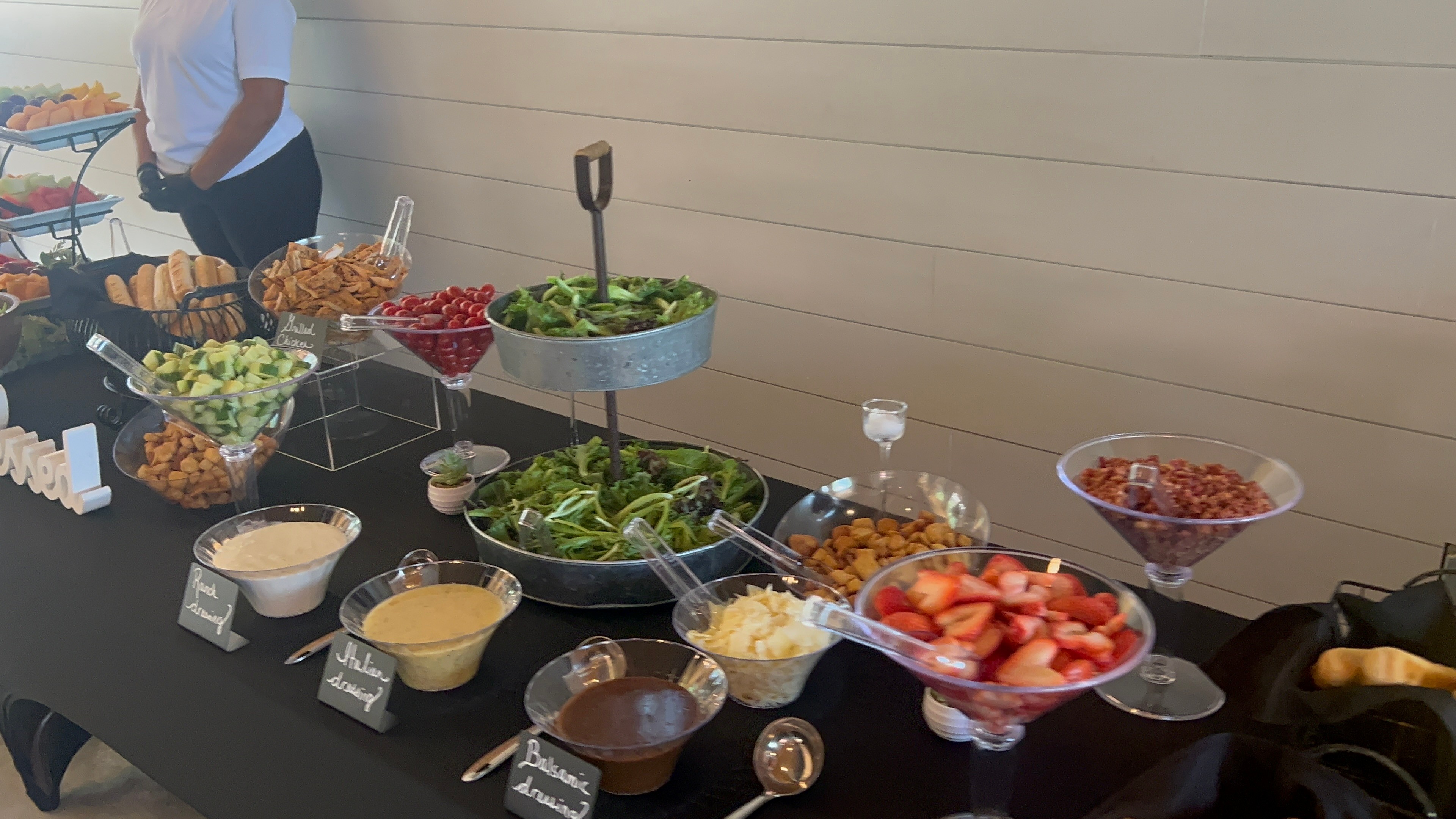 Buffet table with various salads and toppings, person in background, against beige wall.