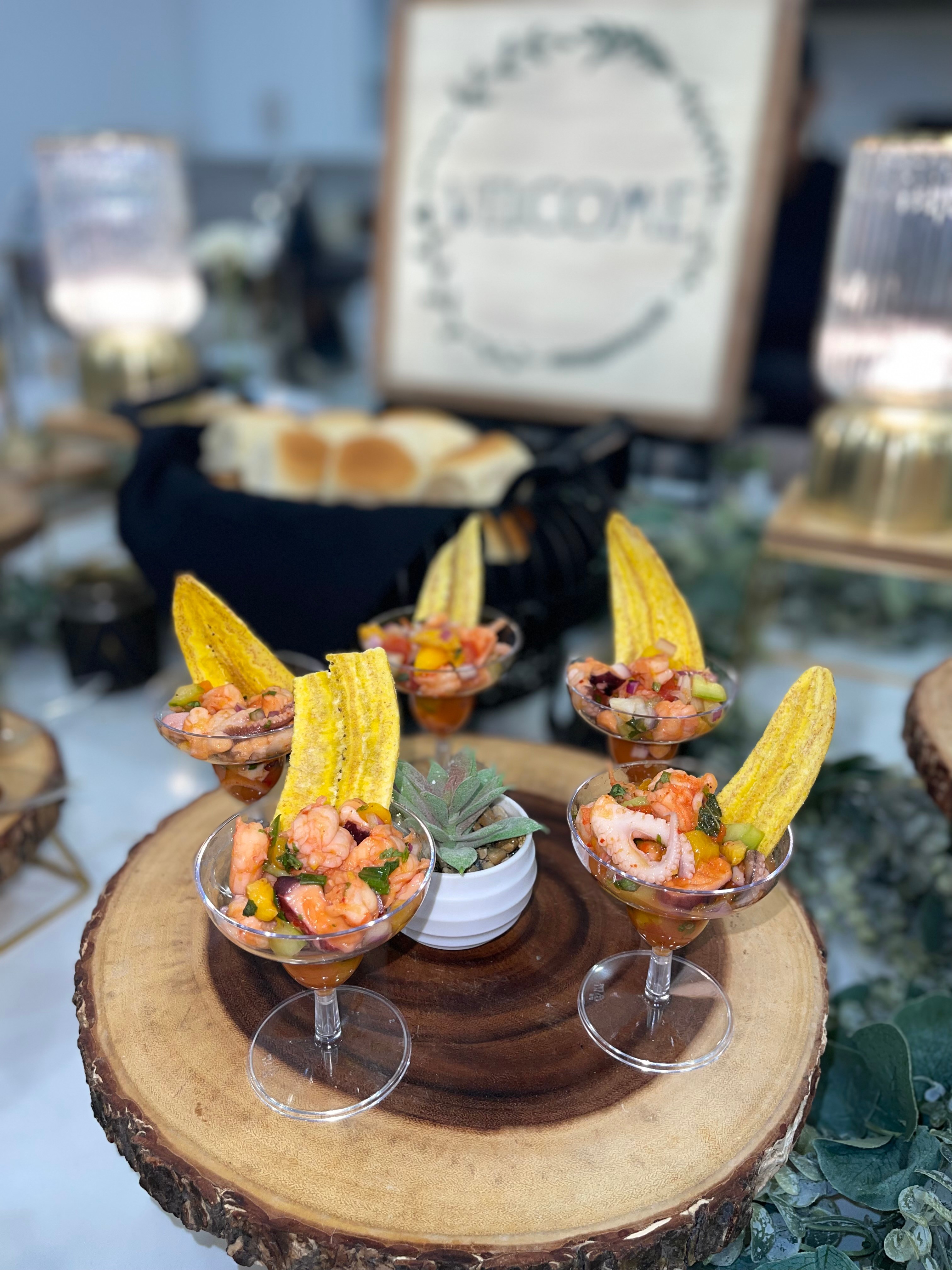 Four small glasses with shrimp and garnishes on a wooden slice, with a blurred background including a framed sign and drinks.