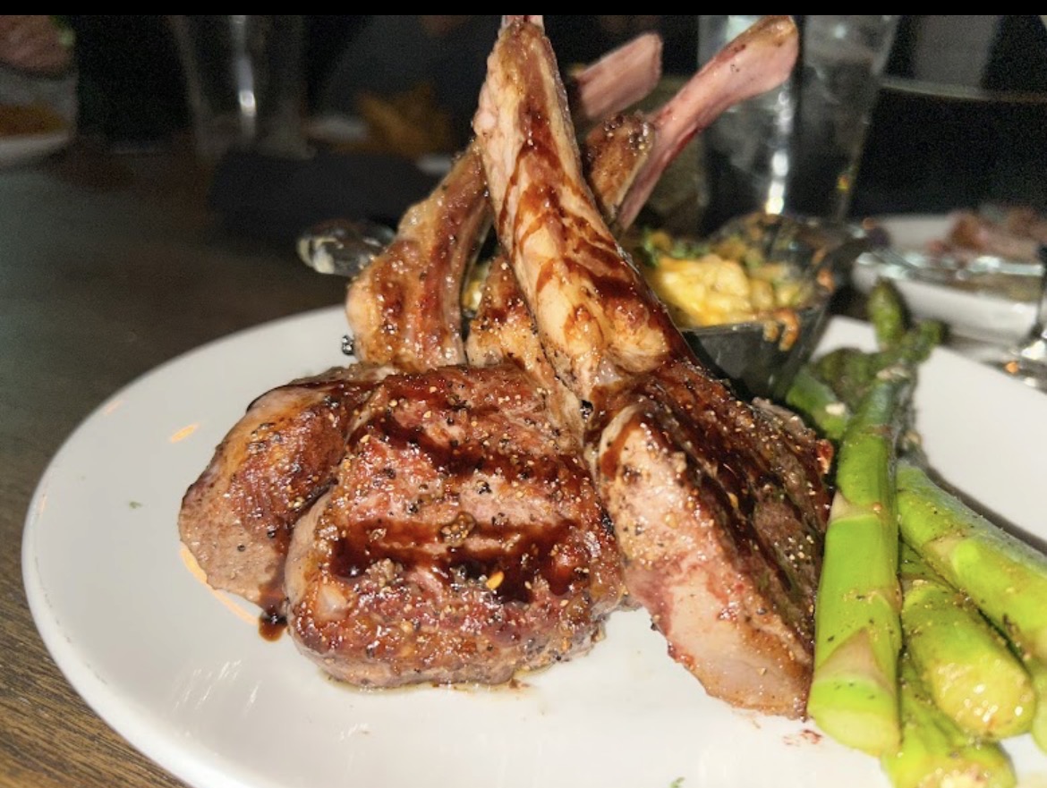 Grilled lamb chops with asparagus on a white plate, with a blurred background of a dining setting.