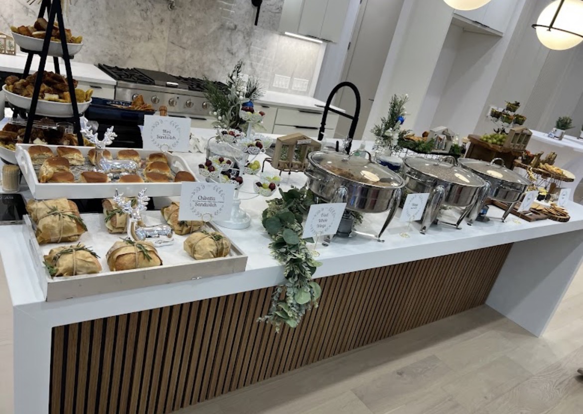 Buffet table with pastries, cereals, and drinks, decorated with greenery and flowers, in a modern kitchen setting.