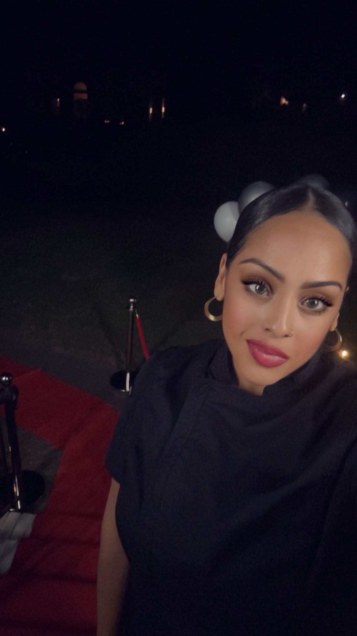 Woman with makeup and hair accessories taking a selfie outdoors at night, with a red carpet and dark background.