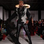Person walking on runway wearing black and white outfit with shoulder embellishments, audience seated on sides, dark ceiling background.