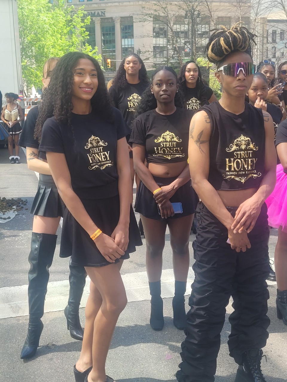 Group of young women standing outdoors, wearing matching black T-shirts with gold text, some with sunglasses, in an urban setting.