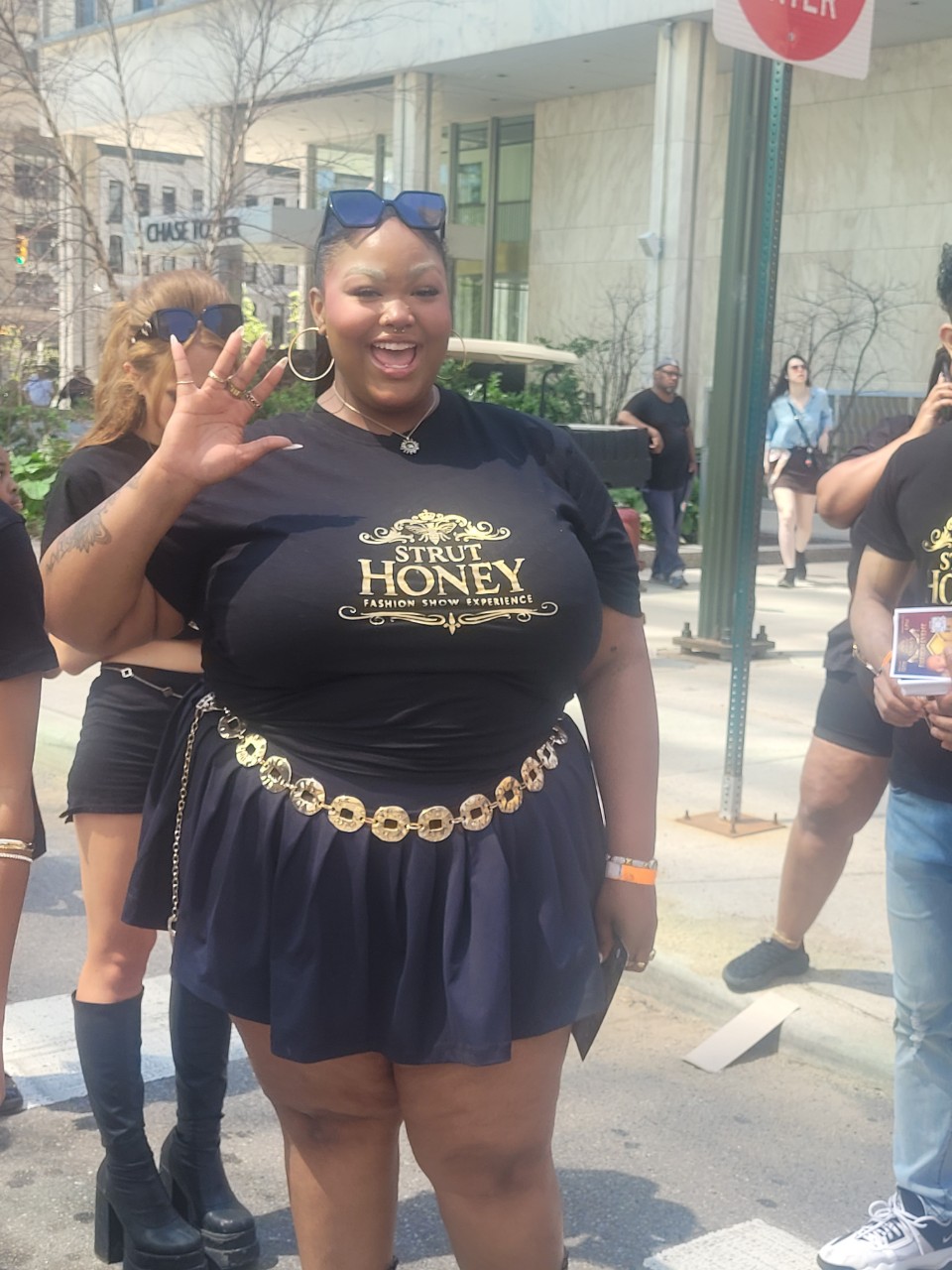 Smiling woman waving outdoors, wearing a black T-shirt, navy skirt, and a gold chain belt, with people and buildings in background.