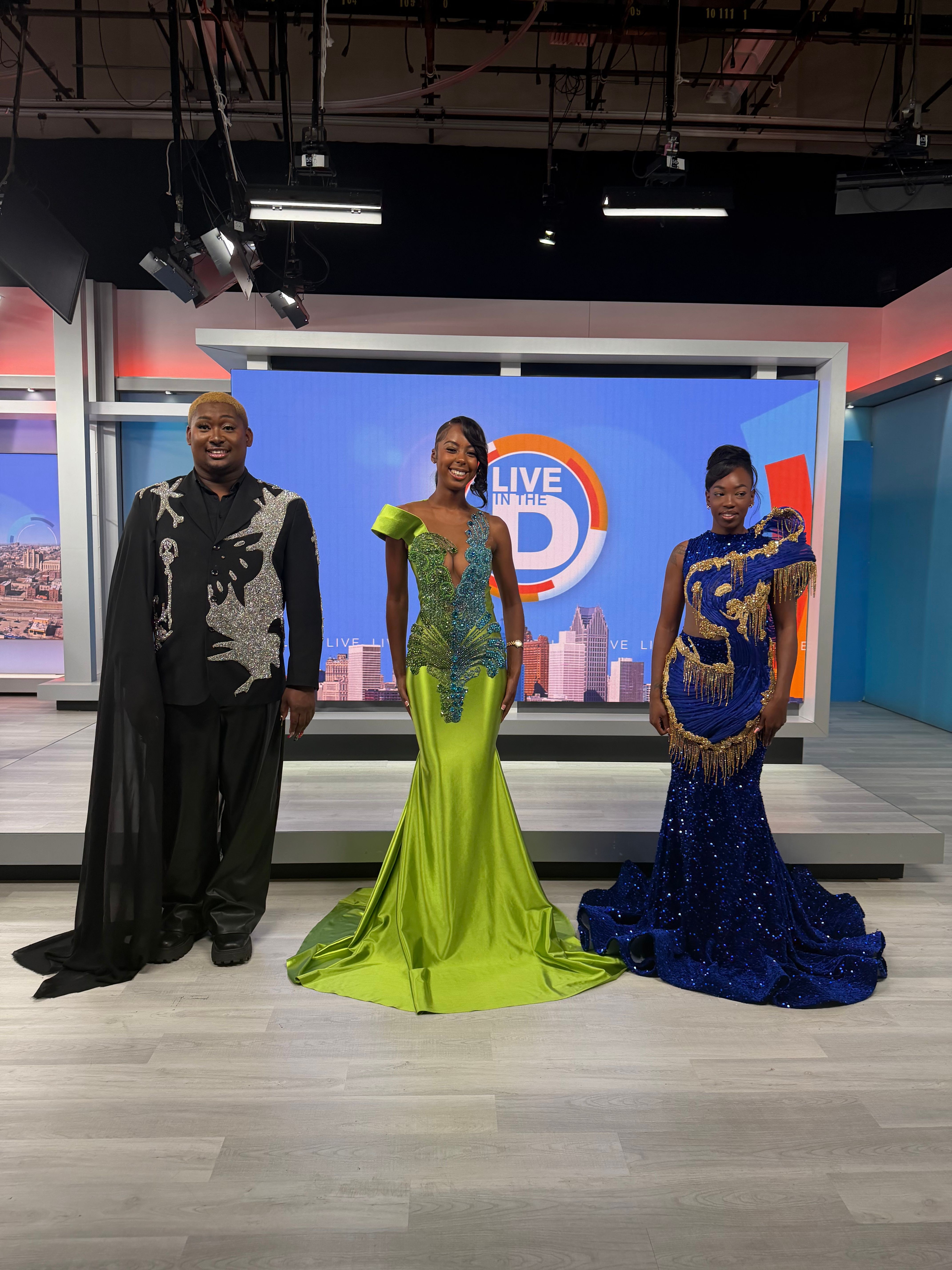 Three people in colorful outfits standing in a TV studio with a screen behind them, two women and one man.