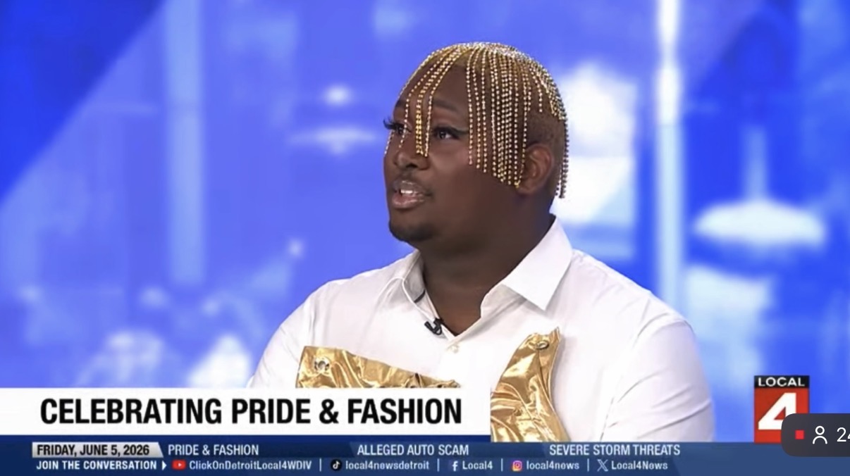 Person with blonde braided hair wearing a white shirt, speaking on a TV show about pride and fashion, with a blue background.