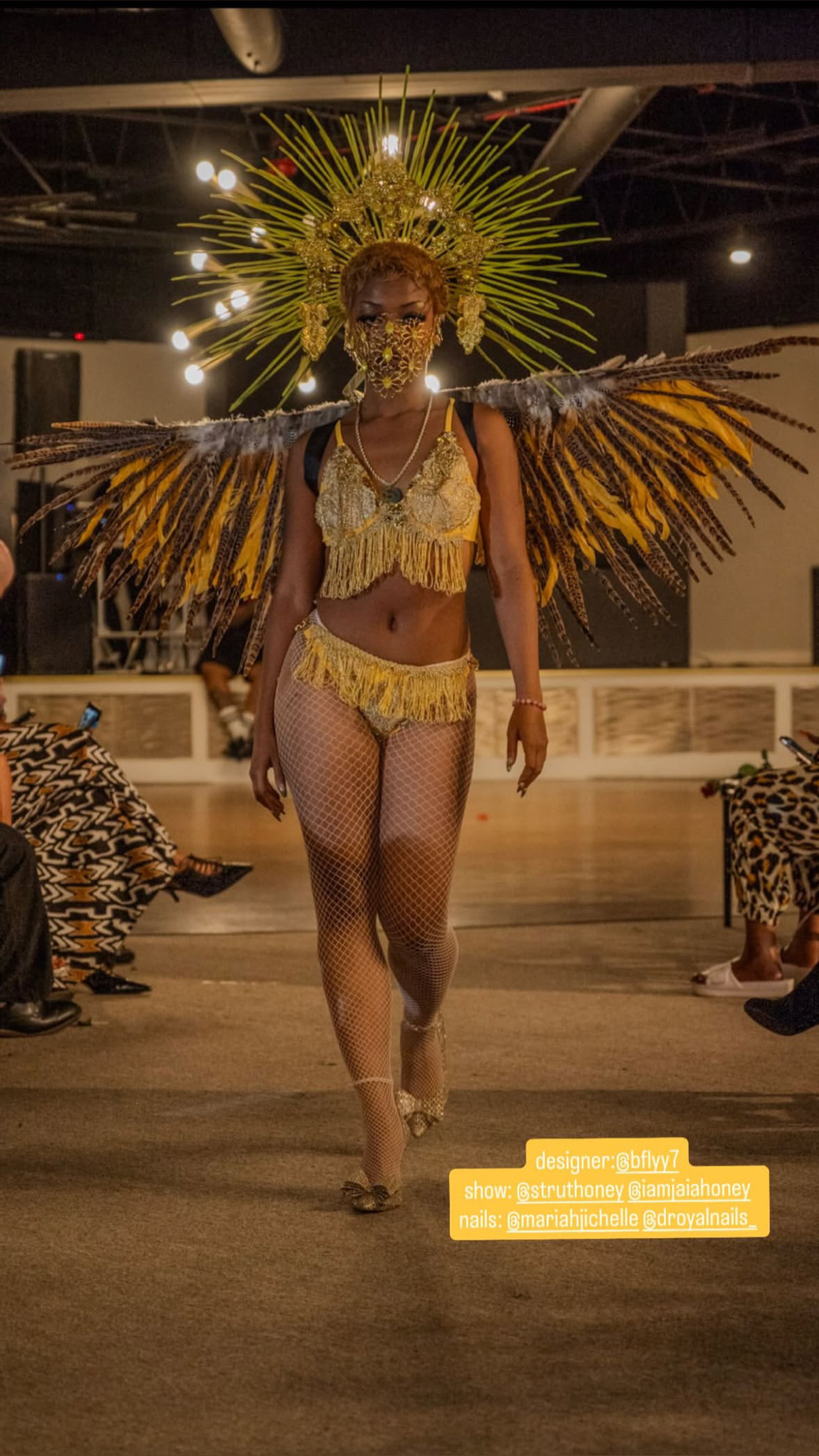 Model walking runway in elaborate costume with large feathered wings and headpiece, audience seated on sides, indoor setting.