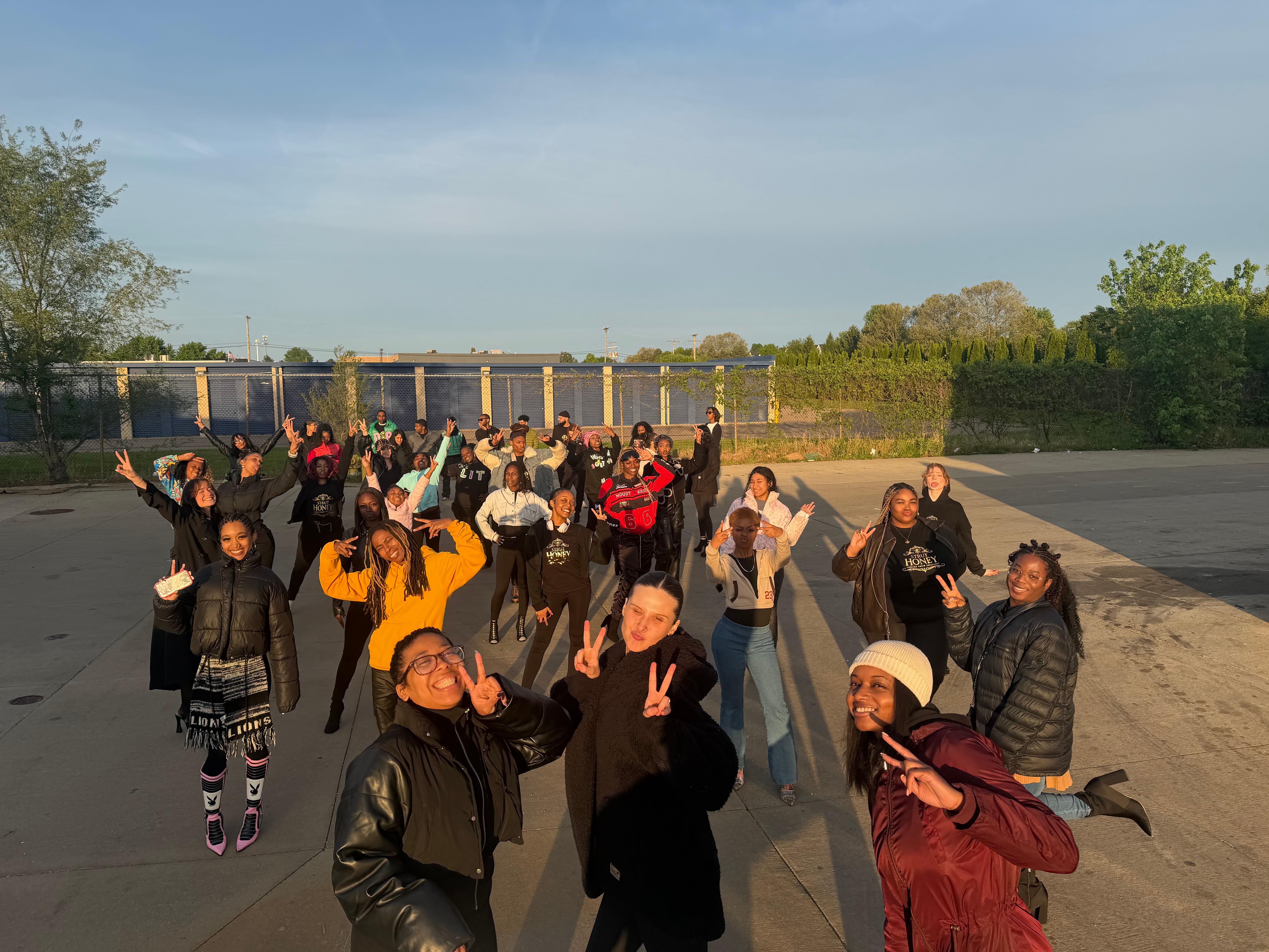 Group of people outdoors, some with arms raised, standing on a paved area with greenery and a fence in the background.