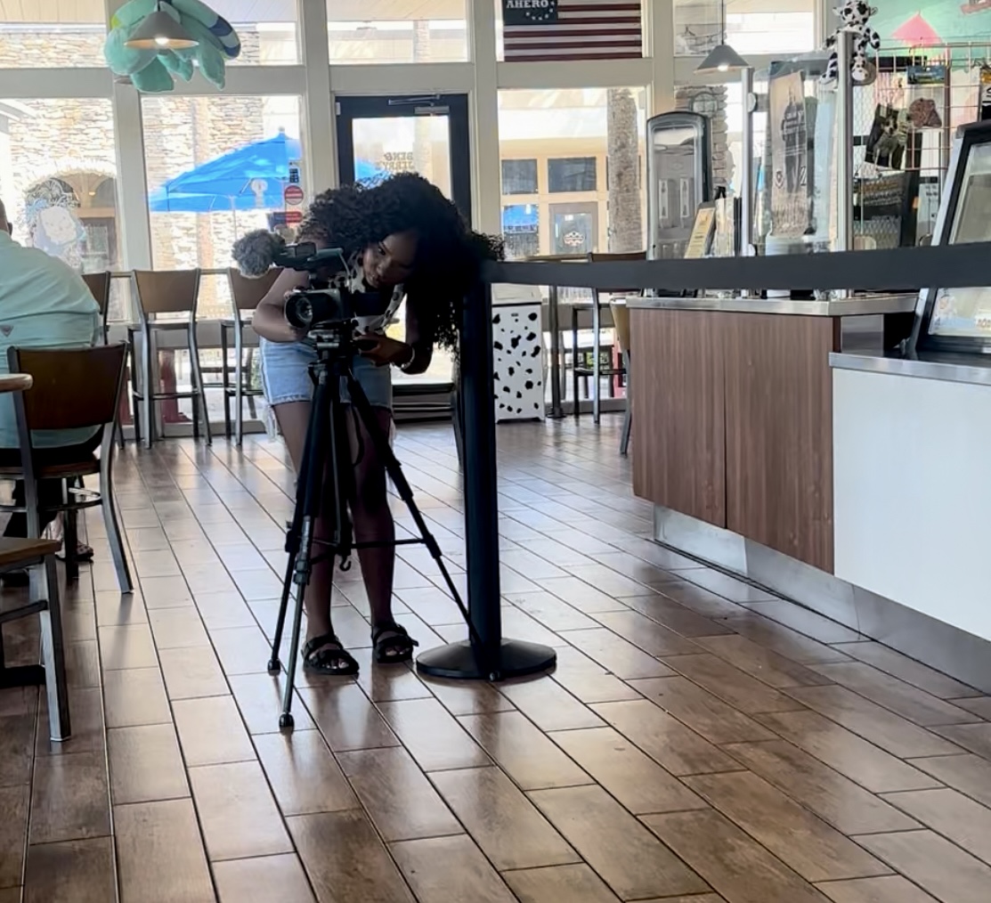 Person with curly hair operating a camera on a tripod inside a cafe or restaurant, with large windows and wooden floor.