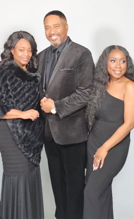 Three people standing together, smiling, dressed in formal black clothing, against a plain white background.