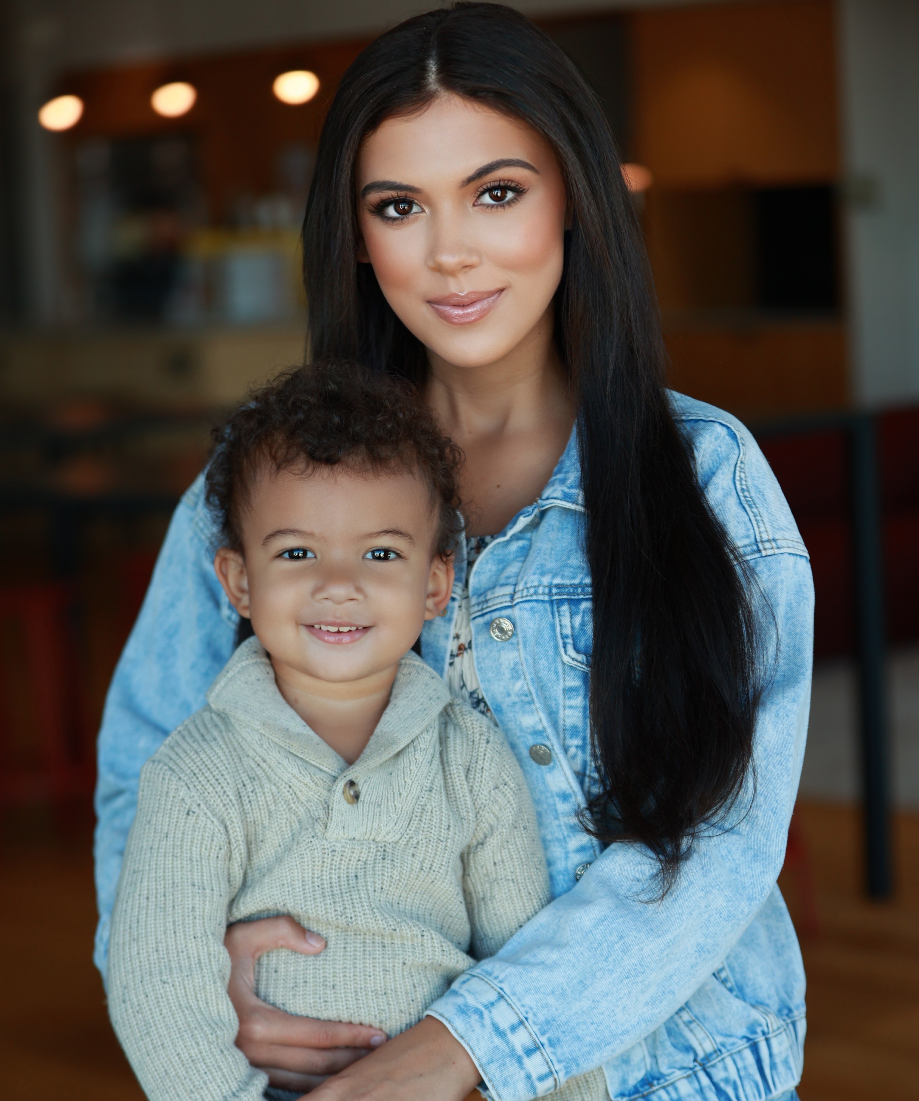 A woman with long black hair and a young child with curly hair smiling, indoors with warm lighting.