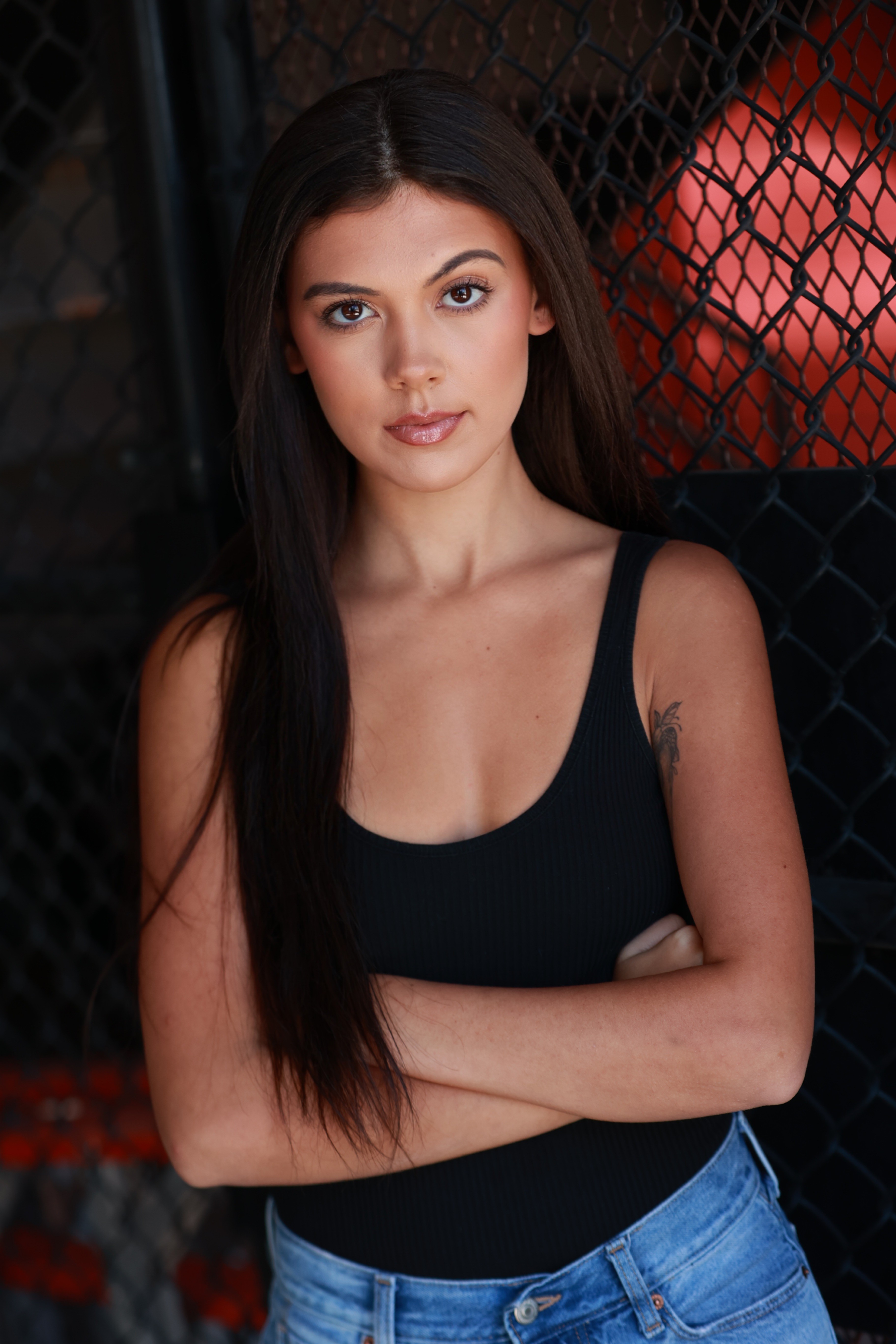 Young woman with long dark hair wearing a black tank top and blue jeans, standing with arms crossed in front of a dark background.