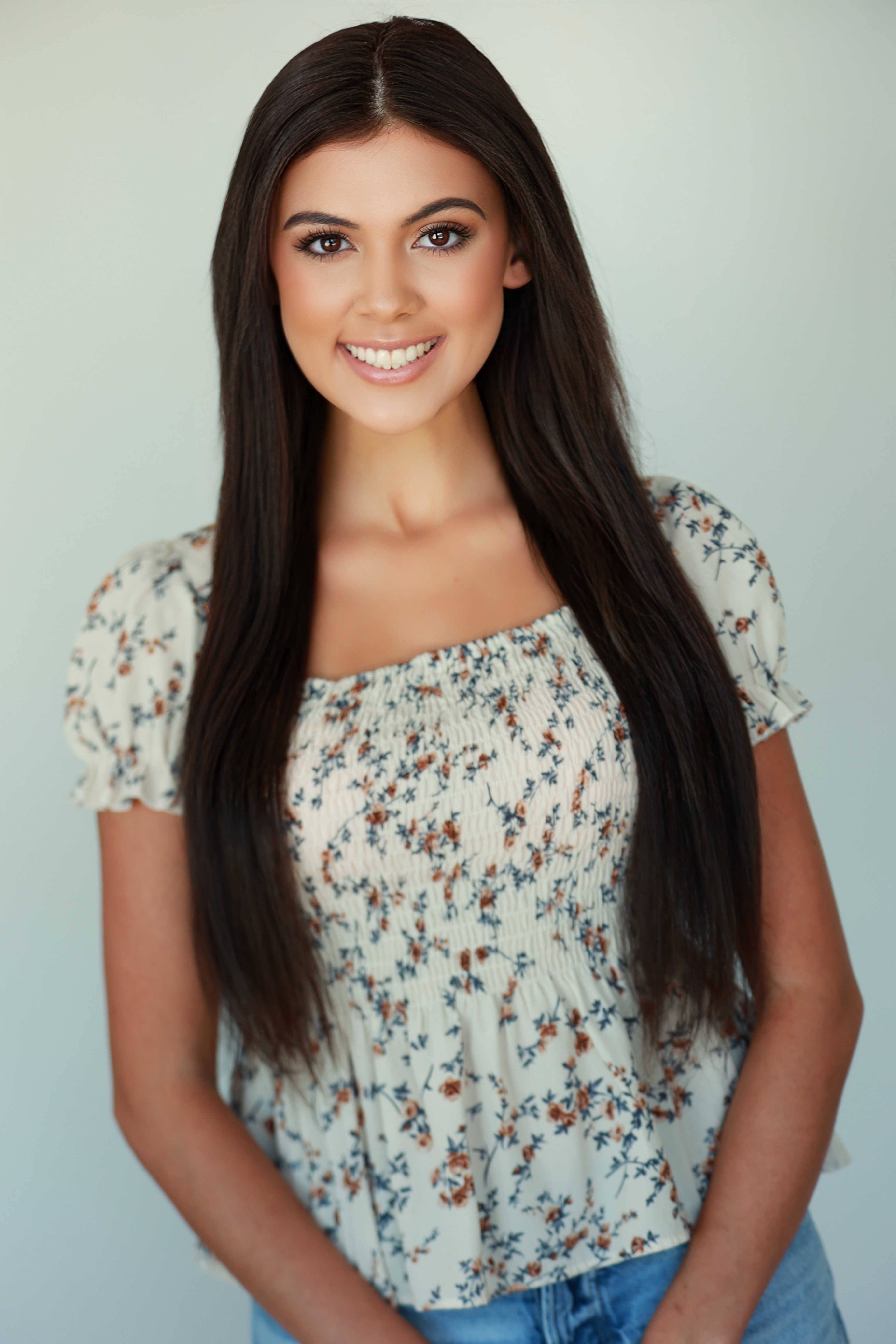 Young woman with long dark hair smiling, wearing a floral top and blue jeans, standing against a light background.