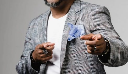 Man with glasses, beard, and dreadlocks wearing a plaid blazer and white shirt, smiling and pointing at the camera.