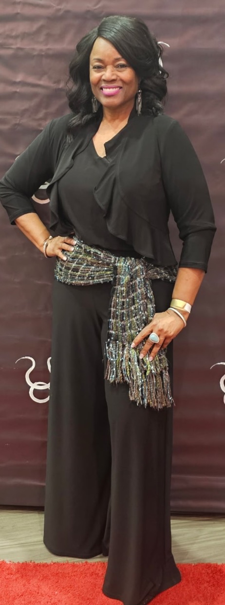 Woman with black hair in a black outfit, smiling, standing with hand on hip, against a dark background.