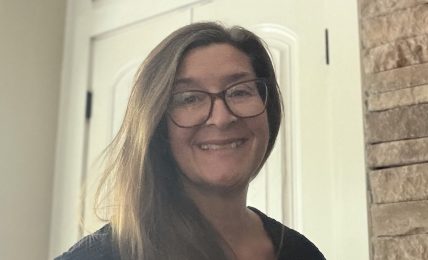 Woman with glasses smiling, standing indoors near a white door and brick wall, wearing a dark top.