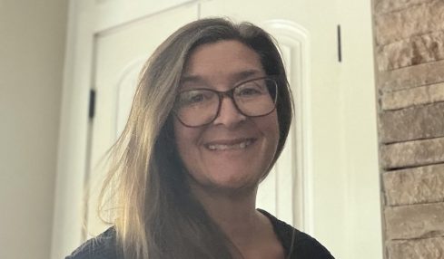 Woman with glasses smiling, standing indoors near a white door and brick wall, wearing a dark top.