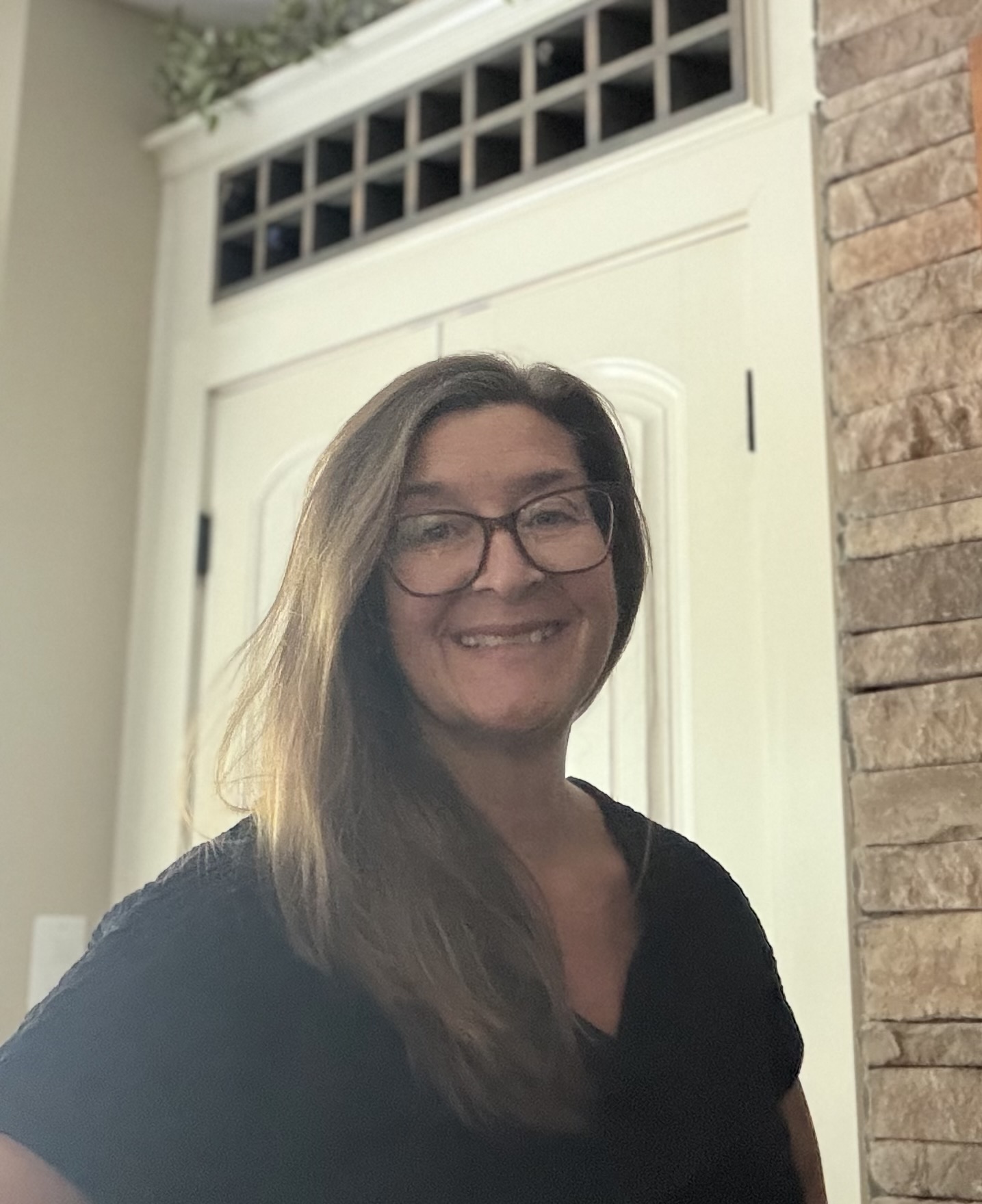 Woman with glasses smiling, standing indoors near a white door and brick wall, wearing a dark top.