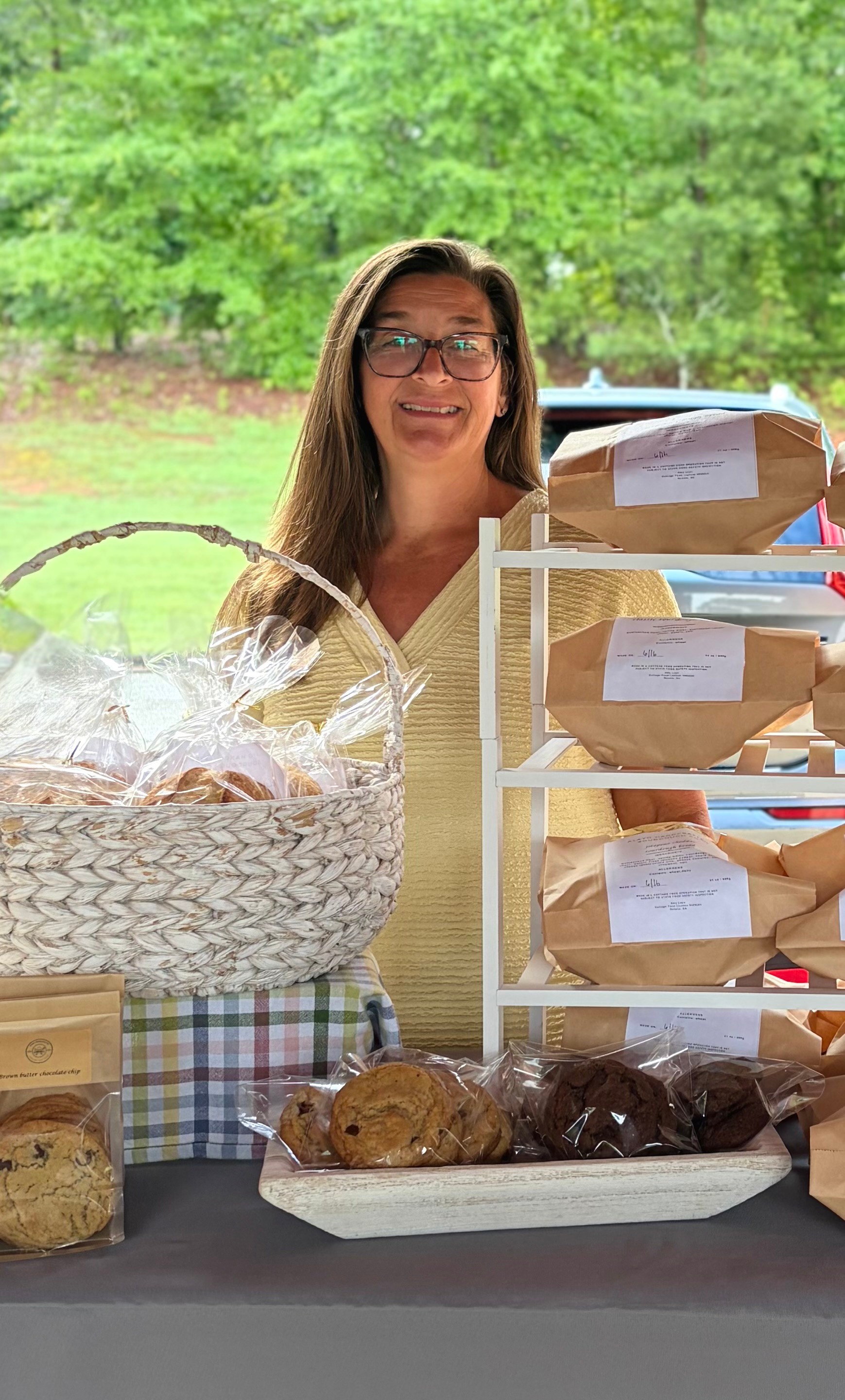 Smiling woman with glasses standing behind a table with baked goods and paper bags, outdoors with green trees in background.