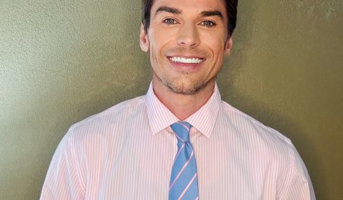 Man with dark hair, wearing a light pink striped shirt and blue striped tie, smiling against a green wall.