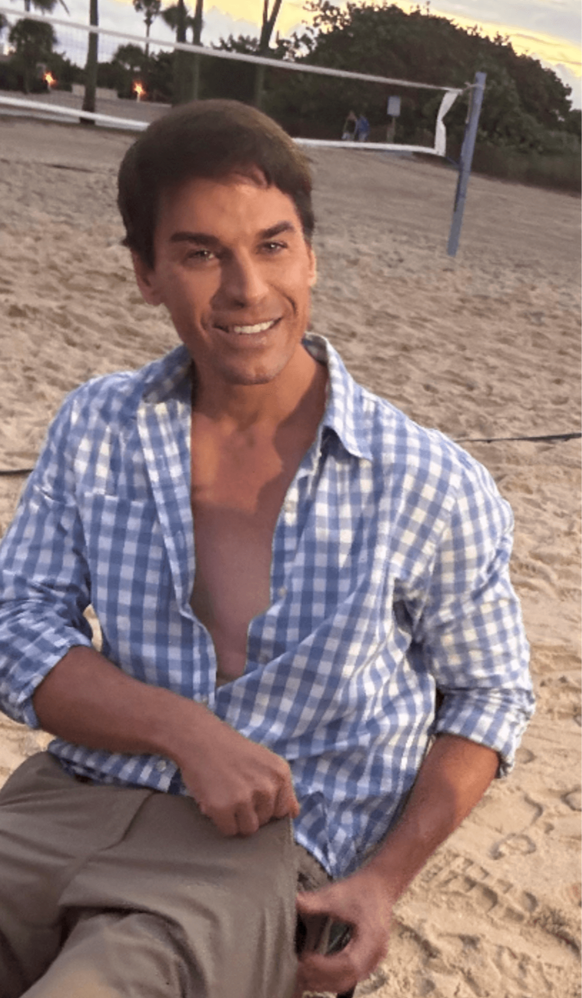 Person sitting on sand at a beach volleyball court, smiling, wearing a blue checkered shirt, with trees in background.