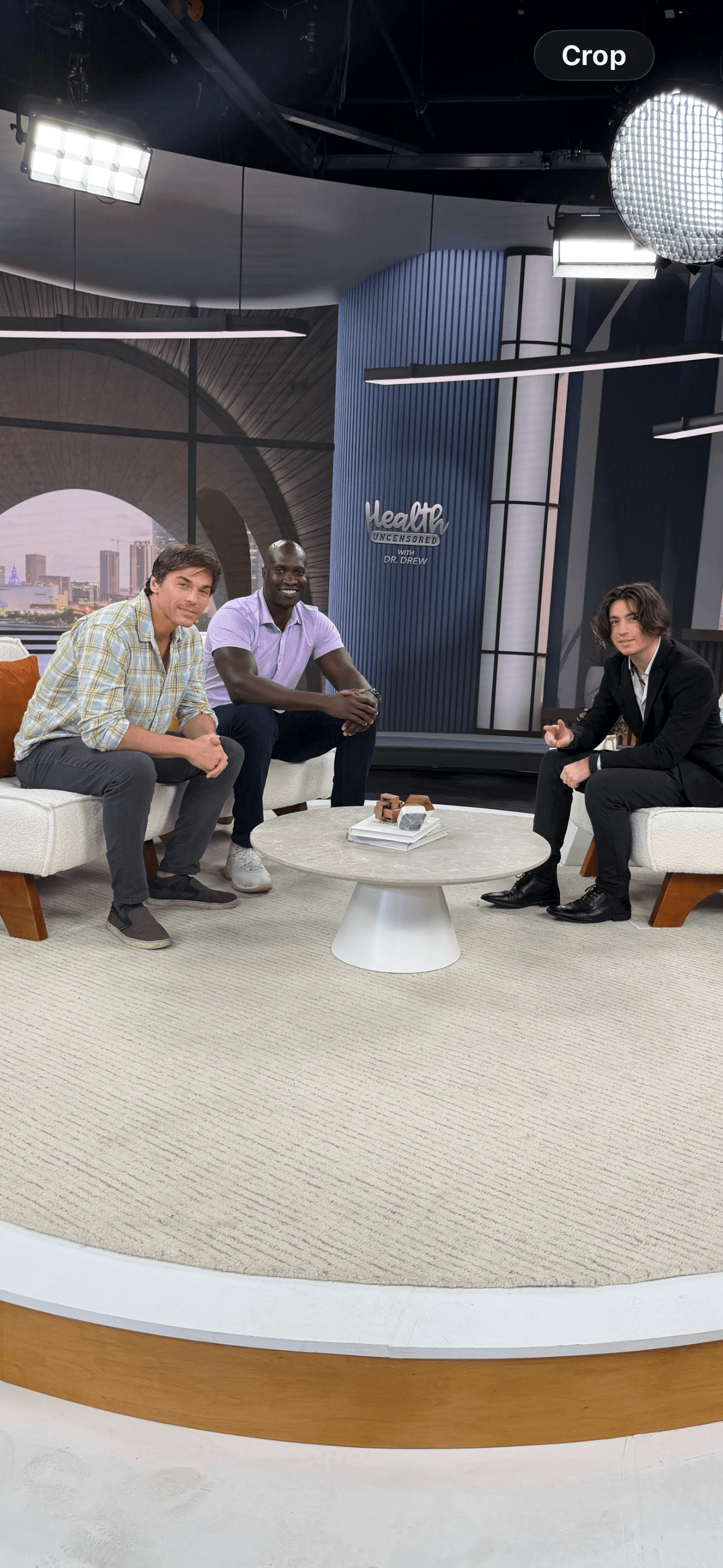 Three people seated in a studio with a cityscape background, discussing around a small table, modern decor, bright lighting.