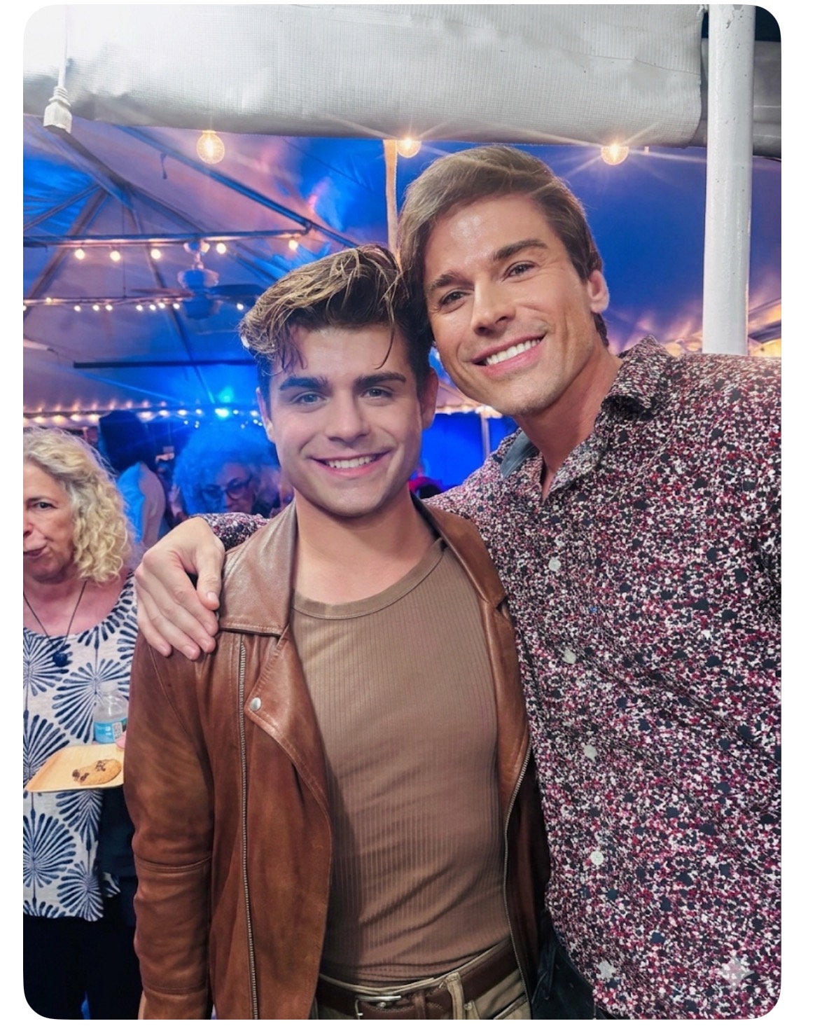 Two young men smiling, one with arm around the other, at a festive event with string lights and people in background.
