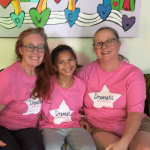 Three smiling people sitting on a couch, wearing pink shirts with 'Dramatic' labels, in front of colorful heart decorations.
