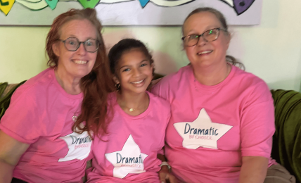 Three smiling people sitting on a couch, wearing pink shirts with 'Dramatic' labels, in front of colorful heart decorations.