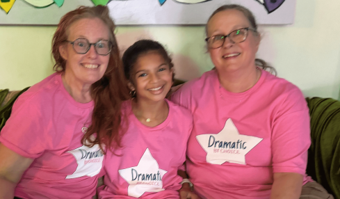 Three smiling people sitting on a couch, wearing pink shirts with 'Dramatic' labels, in front of colorful heart decorations.