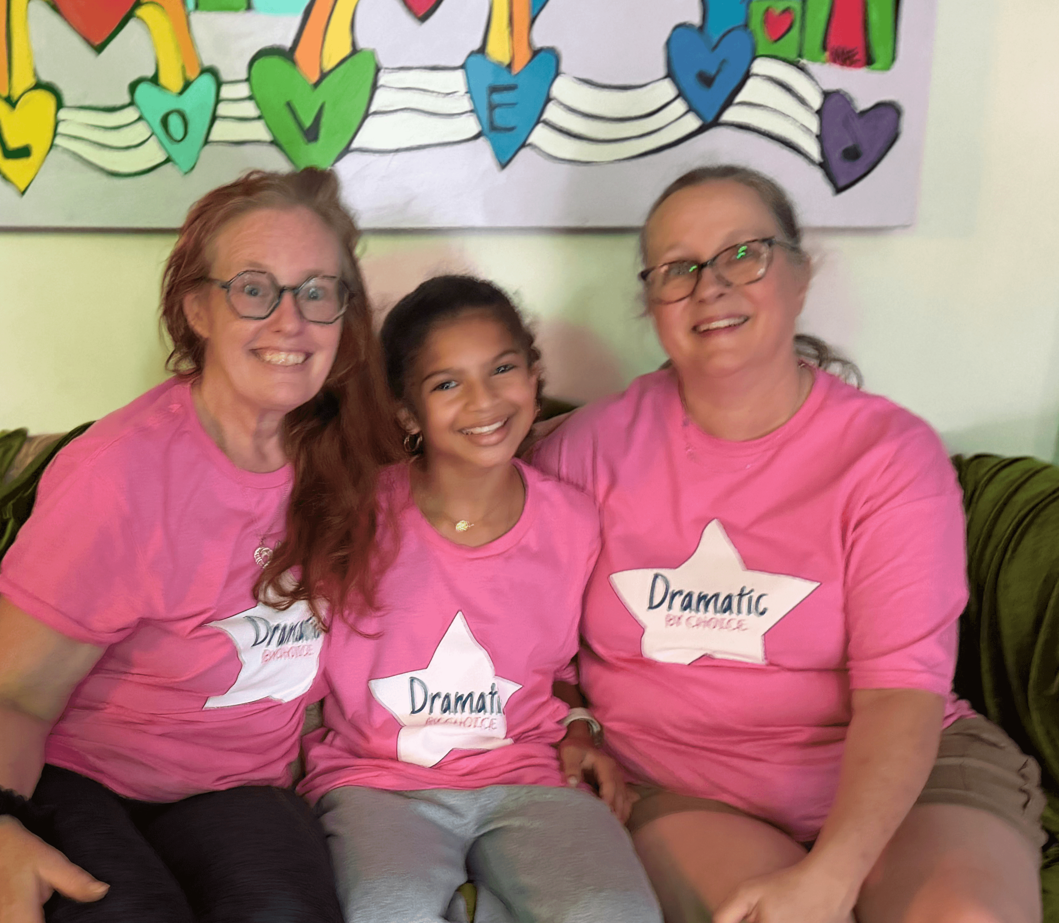 Three smiling people sitting on a couch, wearing pink shirts with 'Dramatic' labels, in front of colorful heart decorations.