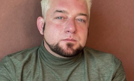 Person with short, bleached hair and a goatee, wearing a green shirt, looking at the camera against a brown background.