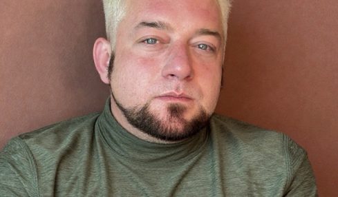 Person with short, bleached hair and a goatee, wearing a green shirt, looking at the camera against a brown background.